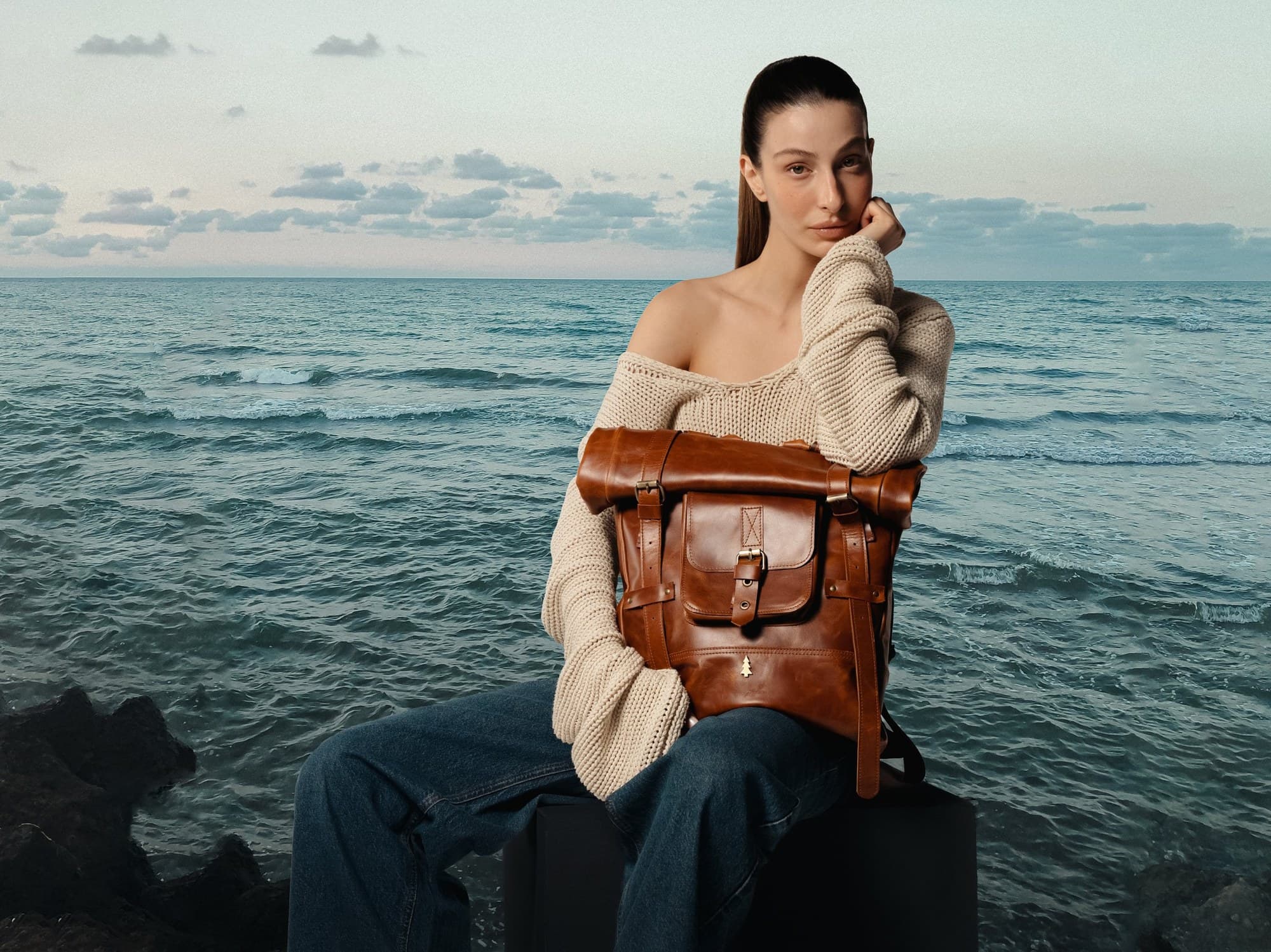 ARAS PINE — model seated by the sea with a cognac roll-top backpack