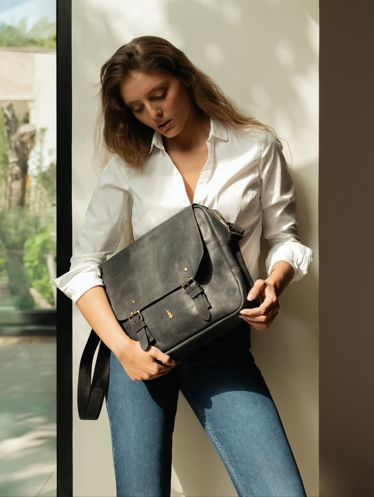 ARAS PINE — model in a white shirt by a sunlit window, holding a distressed graphite full-grain leather messenger bag with the tree emblem