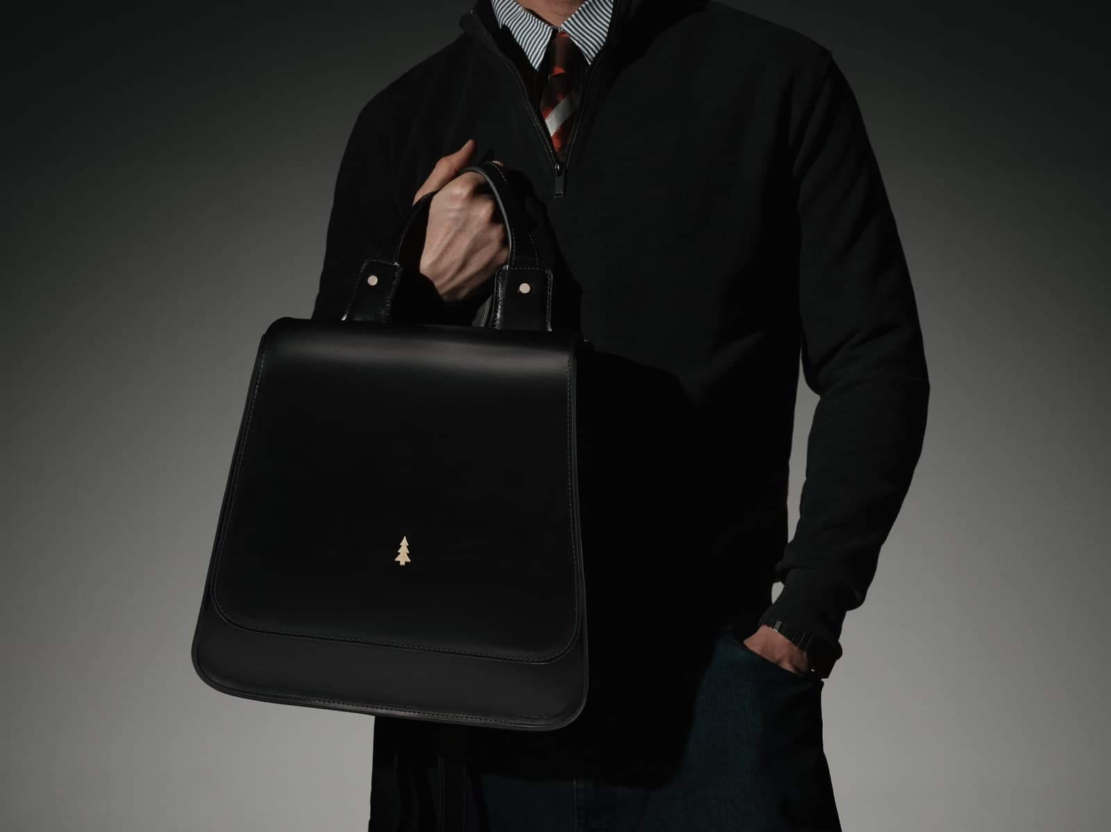 ARAS PINE Valerio Backpack — black — Carried by its top handle at the hip of a man in a black quarter-zip and a burgundy-striped tie. A boardroom piece, boardroom light.