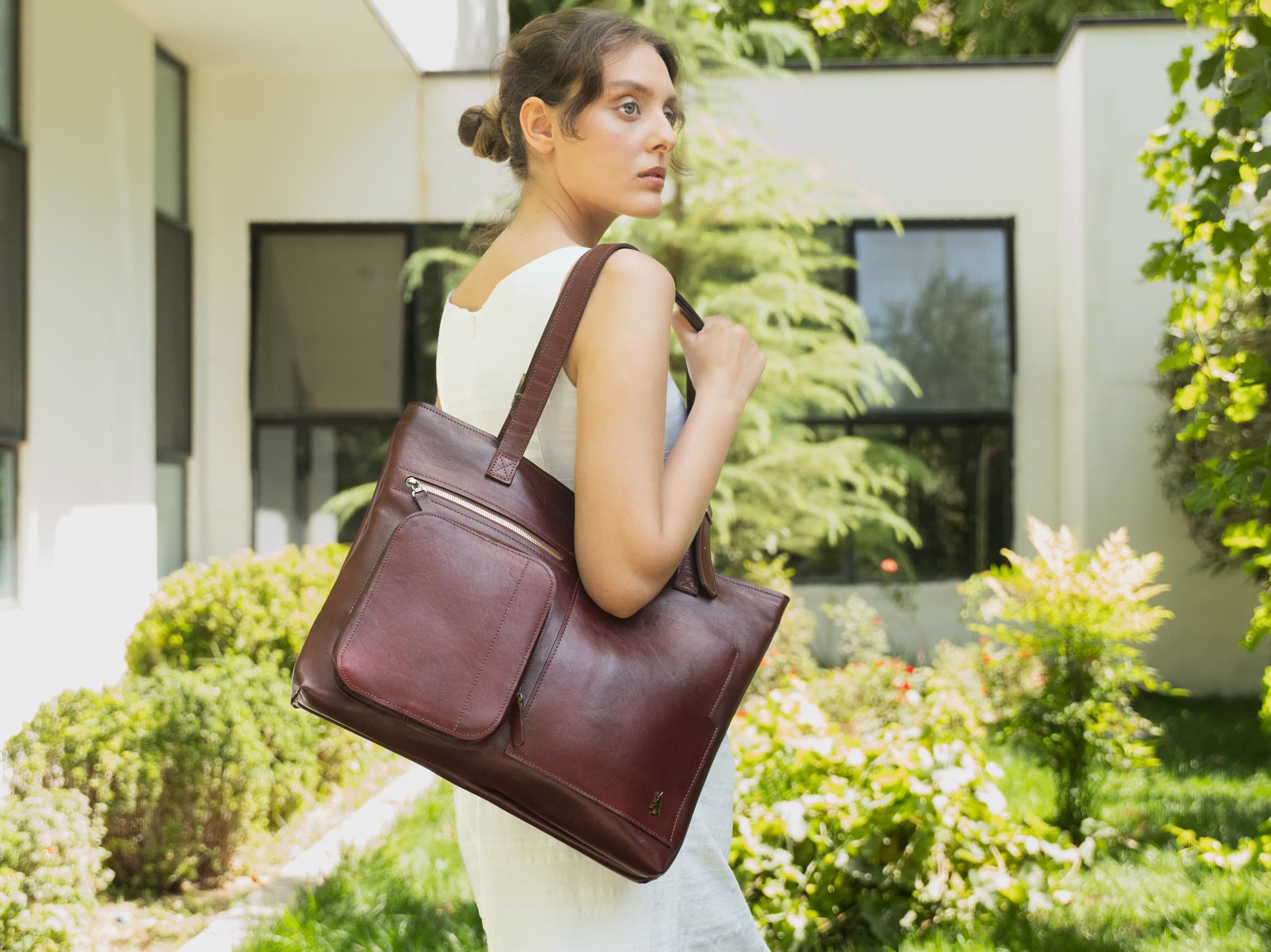 ARAS PINE Lorena Tote Bag — maroon — Structured calfskin at the shoulder, a courtyard in bloom behind. A tote cut for the working week — laptop, notebook, the folded jacket — and quiet enough for the evening it hands off to.