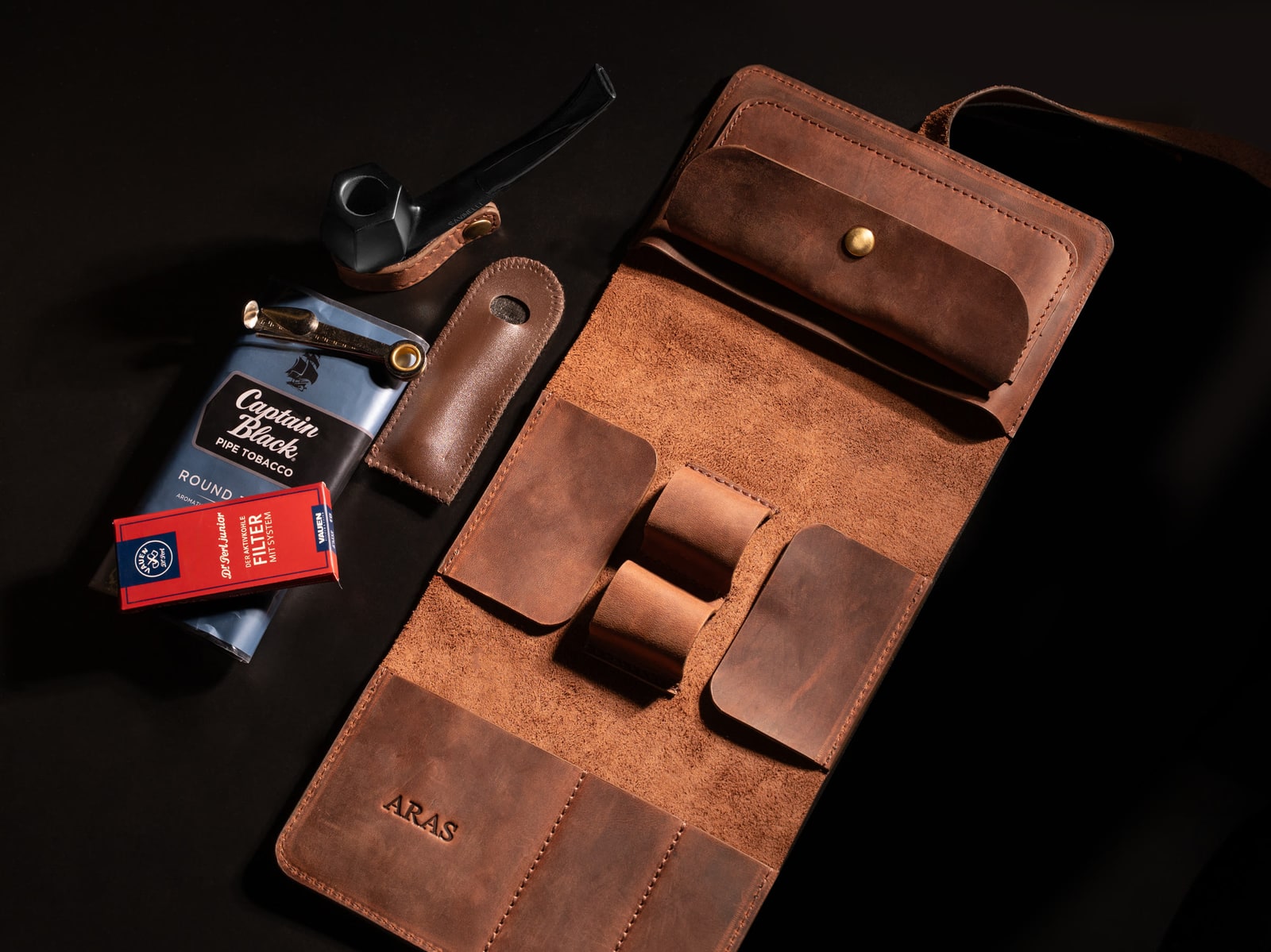ARAS PINE Padrone Pipe Case — unrolled flat-lay on a dark ground with a Savinelli pipe, Captain Black tobacco and a Vauen filter box beside it, ARAS emboss visible on the leather — leather pipe case, handmade in Türkiye
