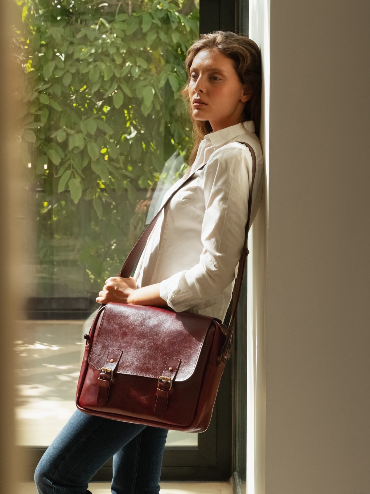 ARAS PINE Bellune Messenger — burgundy, worn cross-body by a model in a white shirt leaning against a villa doorframe with sunlit greenery behind — leather messenger bag, handmade in Türkiye