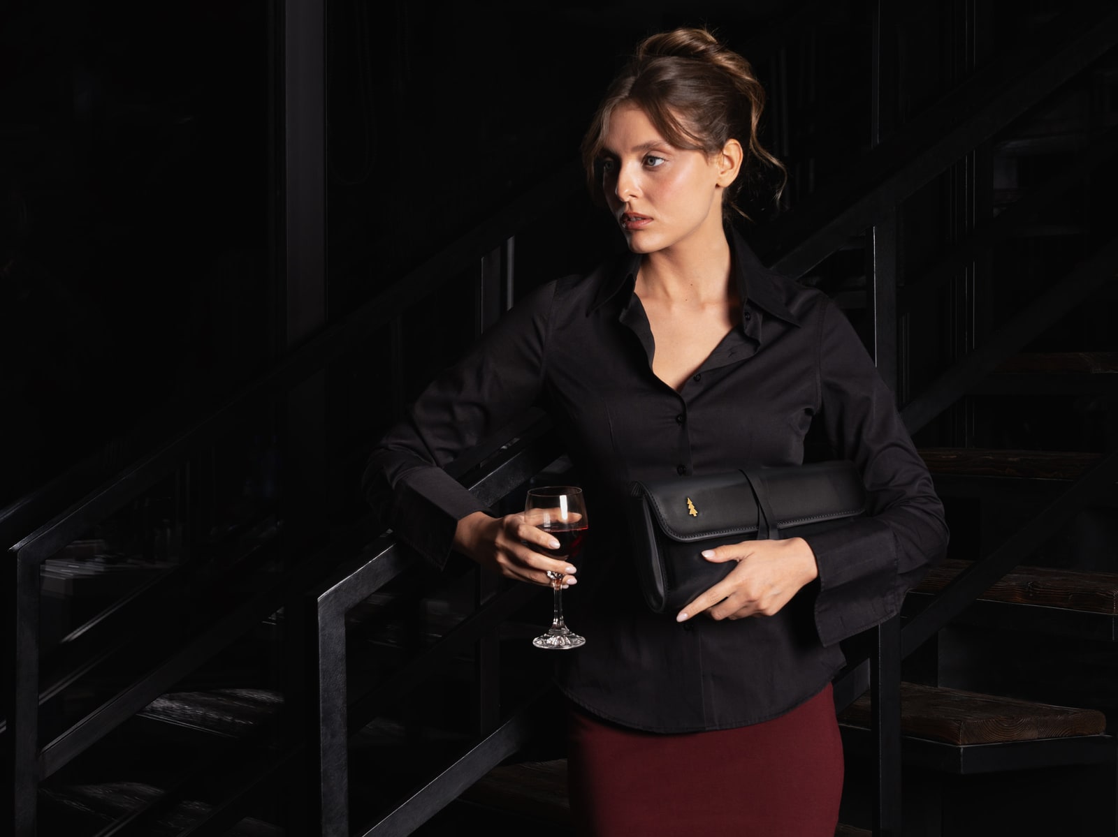 ARAS PINE Corda Clutch — black leather held under the arm of a model on a black steel staircase in a low-lit bar, wine glass in the other hand — leather clutch, handmade in Türkiye
