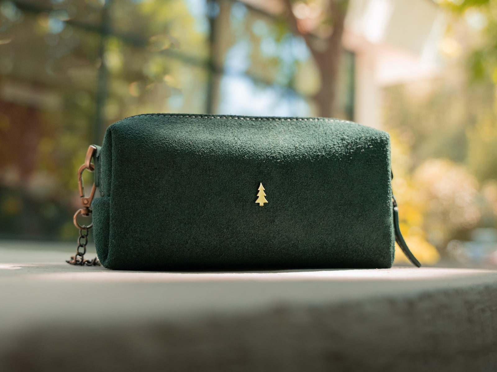 ARAS PINE Ivrea Crossbody — green suede, brass emblem catching afternoon light on a stone ledge with autumn bokeh behind — leather crossbody bag, handmade in Türkiye