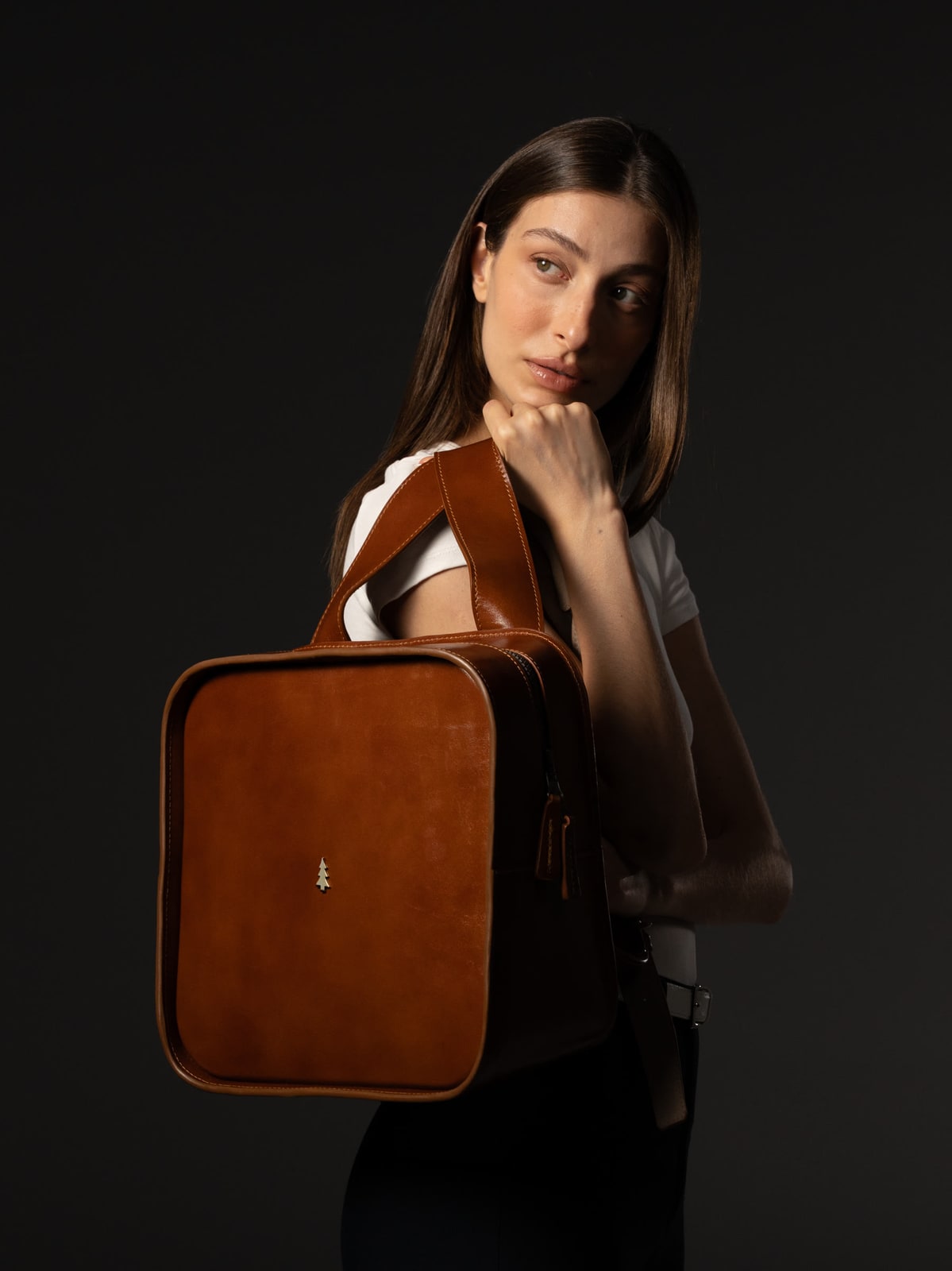 ARAS PINE Montel Backpack in cognac full-grain calfskin with the brass pine icon on the front panel — slung one-shouldered by a model in a white tee under a hard side-spot against a black studio backdrop