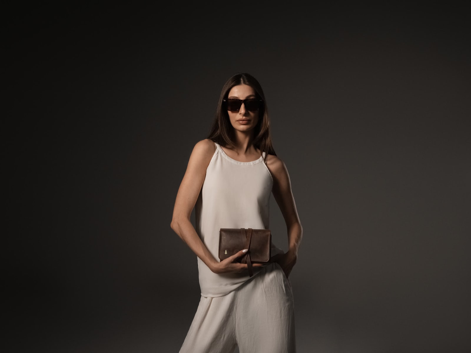 ARAS PINE Padrone Pipe Case in brown roll-and-tie leather with the brass pine icon on the flap — held at the waist of a model in sunglasses, cream silk cami and cream palazzo trousers against a deep grey studio backdrop