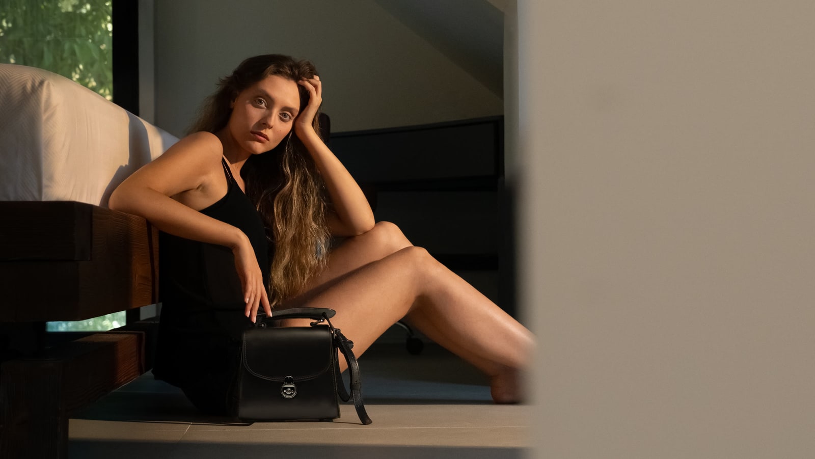 ARAS PINE Top-Handle Handbag in black leather with brass push-lock — set beside a model in a black slip dress seated on the floor of a sunlit villa bedroom in Doha